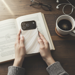 White silk-and-gold wide-camera case held above an open book and coffee #iphone-17-pro