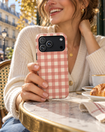 Woman in a cream cardigan holding a pink Prairie Gingham phone case at an outdoor café