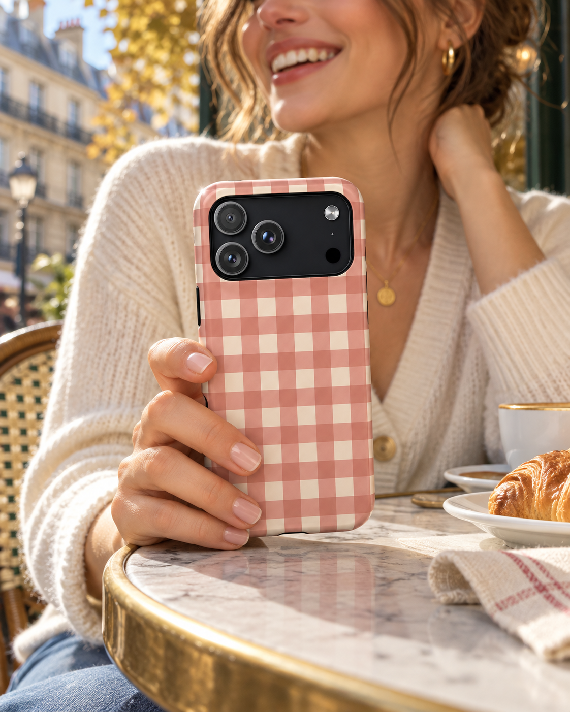 Woman in a cream cardigan holding a pink Prairie Gingham phone case at an outdoor café